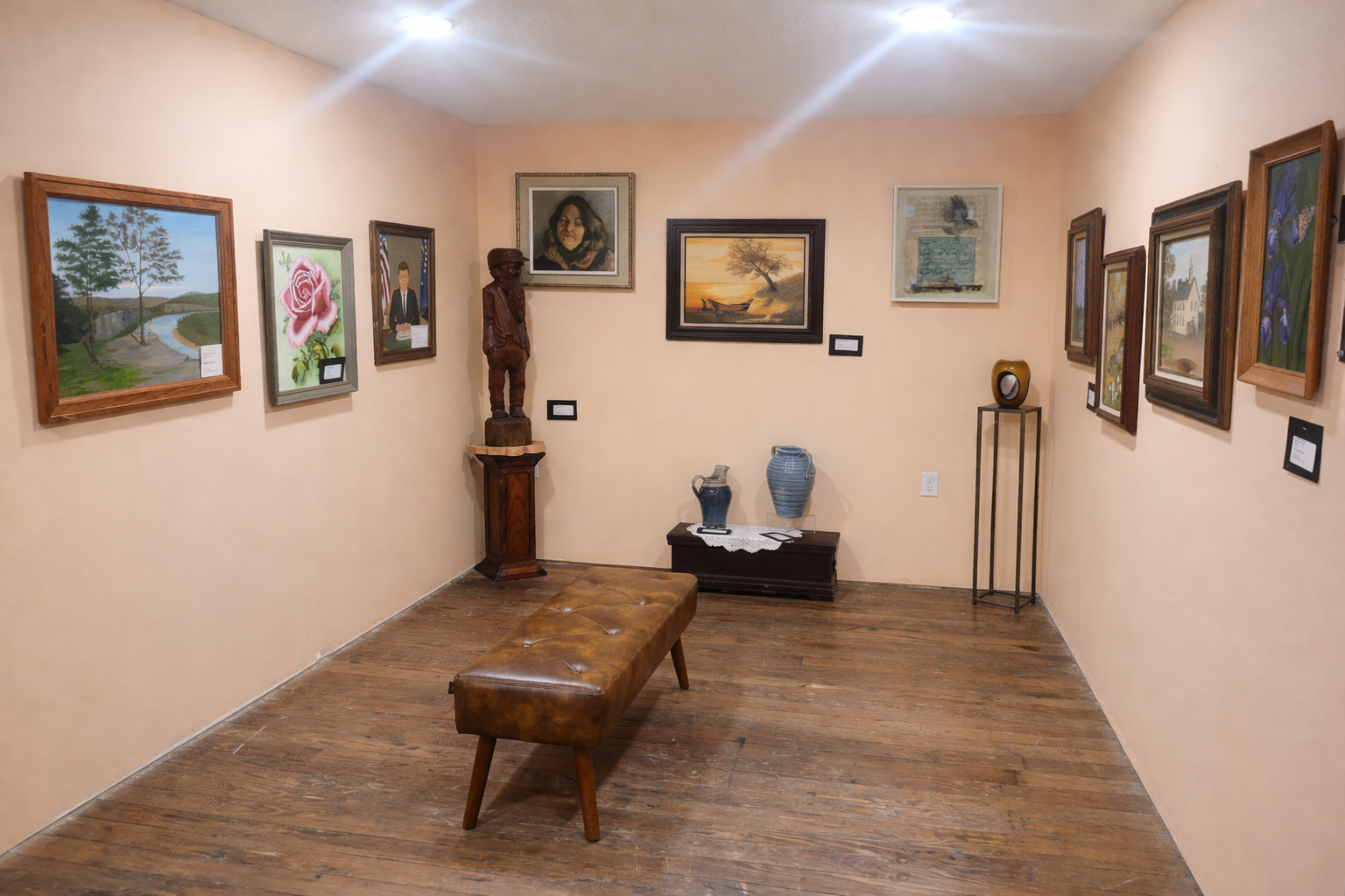 Fine art gallery room with framed paintings, sculpture, pottery, bench seating, wood flooring, and gallery lighting