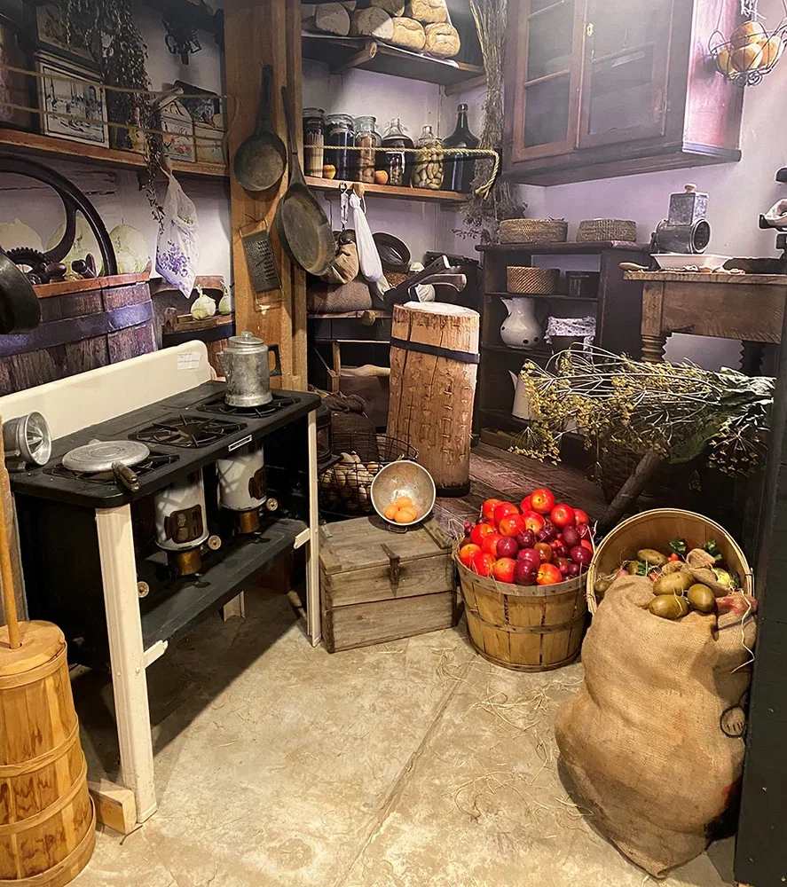 Living-history domestic interior with stove, cookware, baskets, preserved goods, apples, potatoes, and household items
