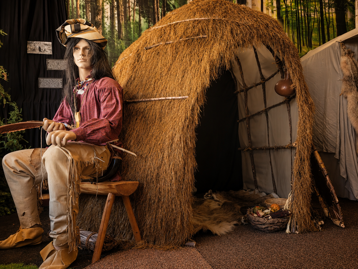 Native History exhibit with a hand-built wickiup, seated figure, forest backdrop, hides, tools, and basket details