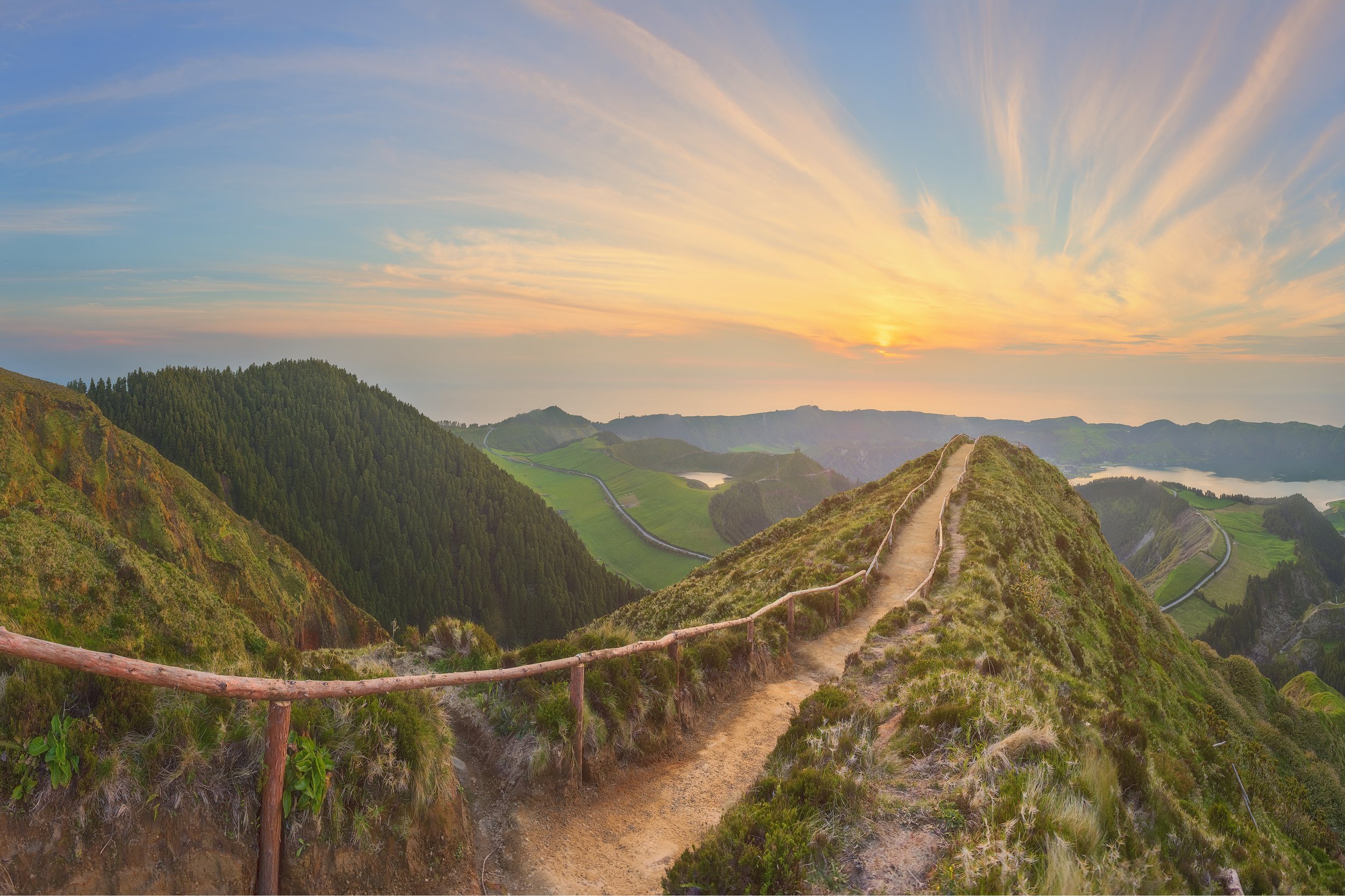 Scenic view of a winding path leading through lush green hills with a sunset sky in the background.