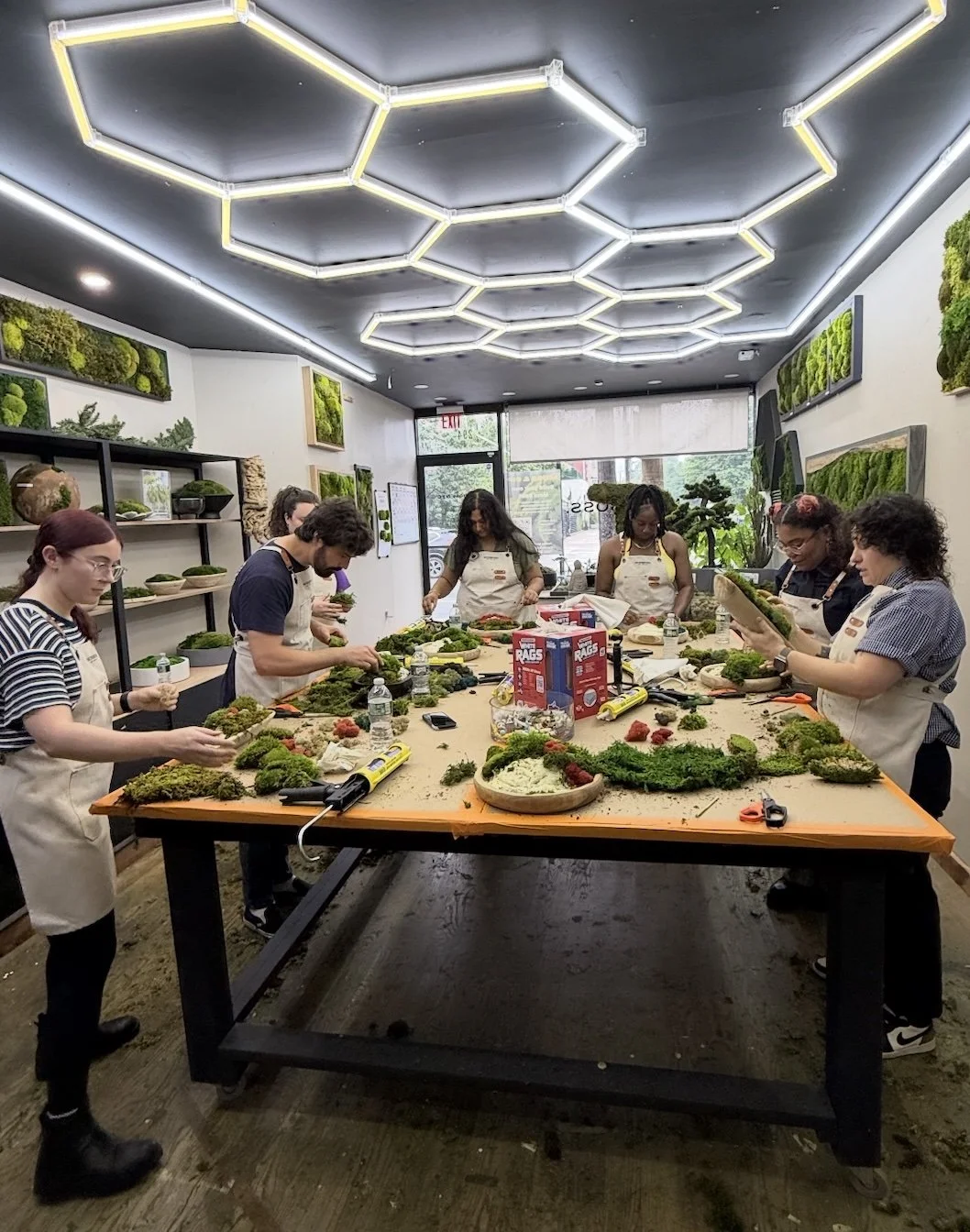 A full class gathered around the work table under the honeycomb light fixture at MossBoss NYC