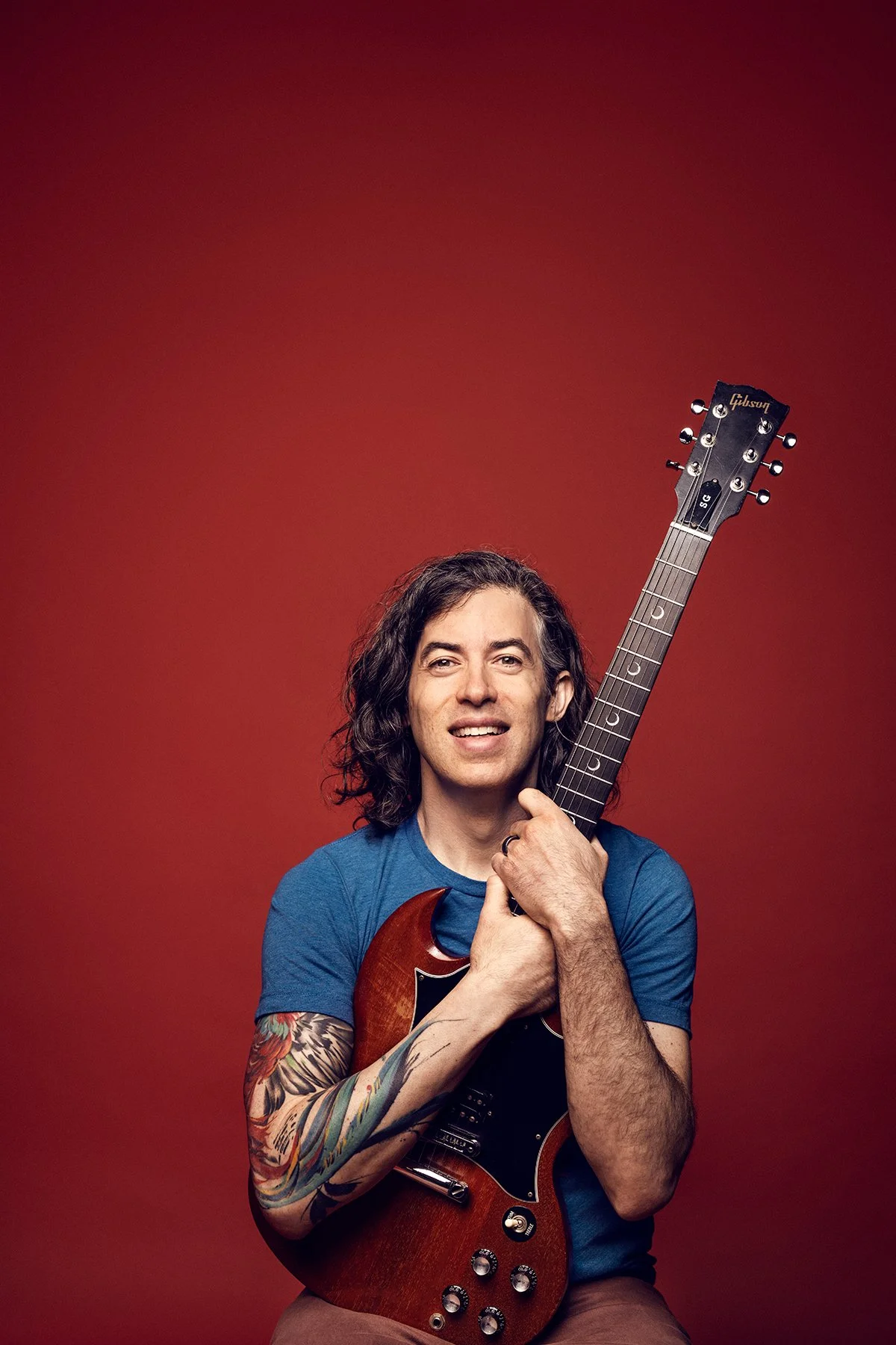 Dave Reminick smiling, holding a cherry-red Gibson SG upright against a deep red background