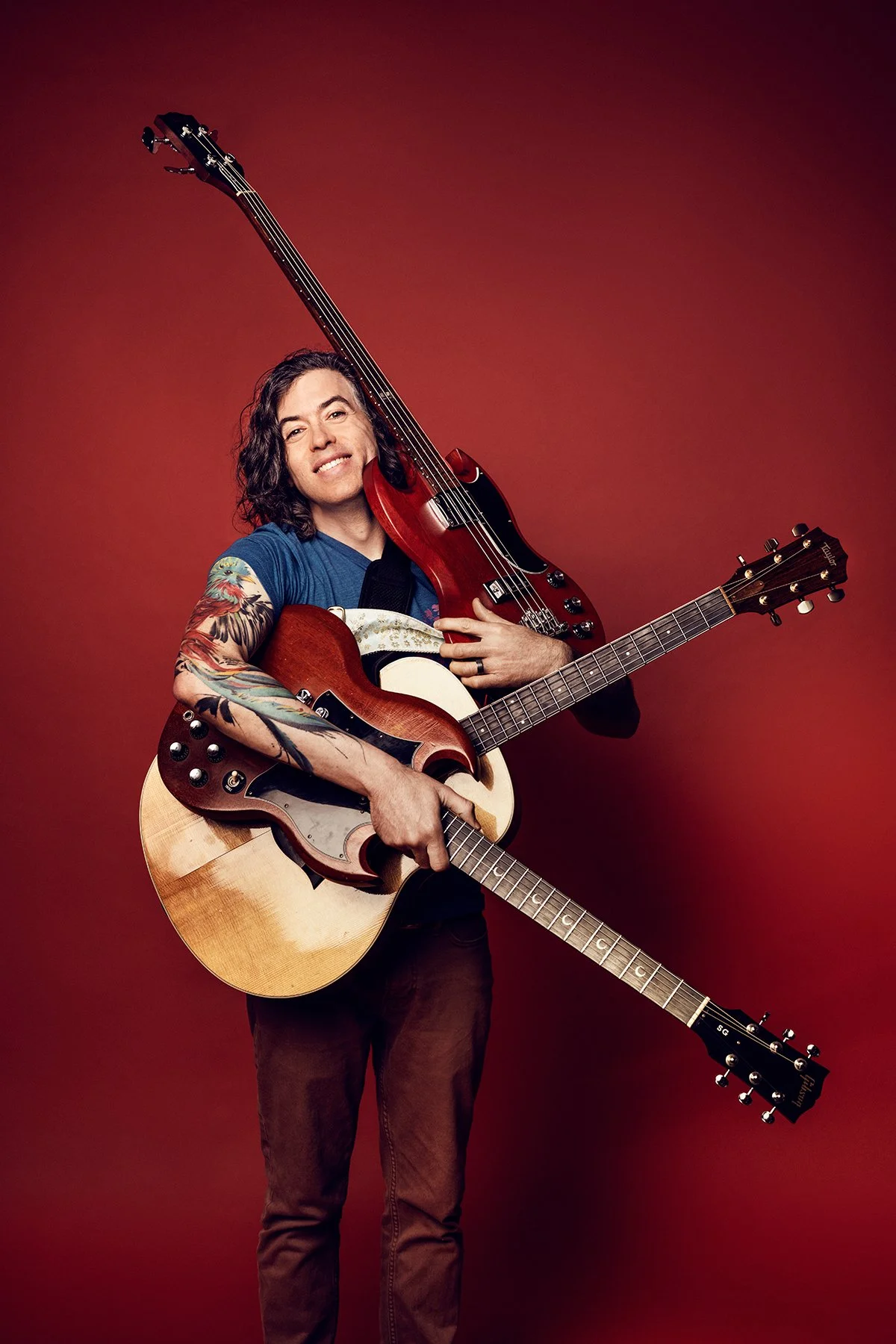 Dave Reminick against a red background holding three guitars at once: an acoustic, an SG and an SG bass
