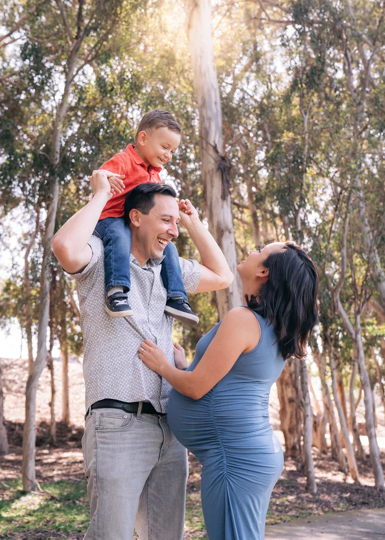 Expecting family in a eucalyptus grove, father holding their young son on his shoulders and mother in a fitted blue maternity gown