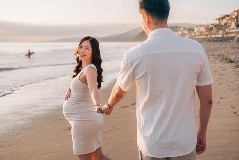 Expecting couple walking barefoot at the water's edge at sunset, the mother in a fitted cream tank dress looking back over her shoulder and her partner in a cream linen shirt, a surfer in the shallows behind them