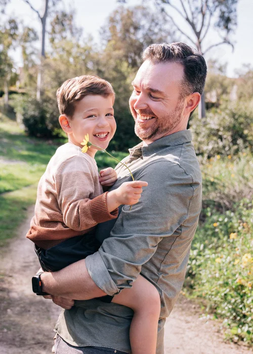 Dad holding his son, who grins back over his shoulder with a yellow daisy in hand
