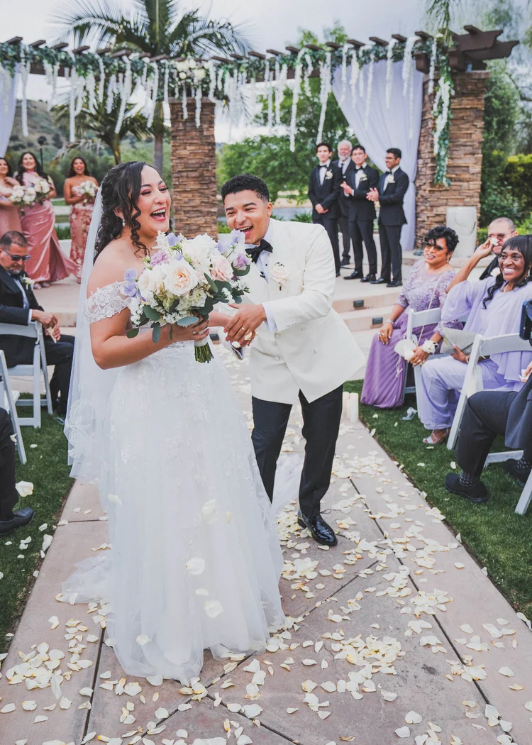 A bride and groom laughing together as they walk up a petal-covered aisle past smiling guests