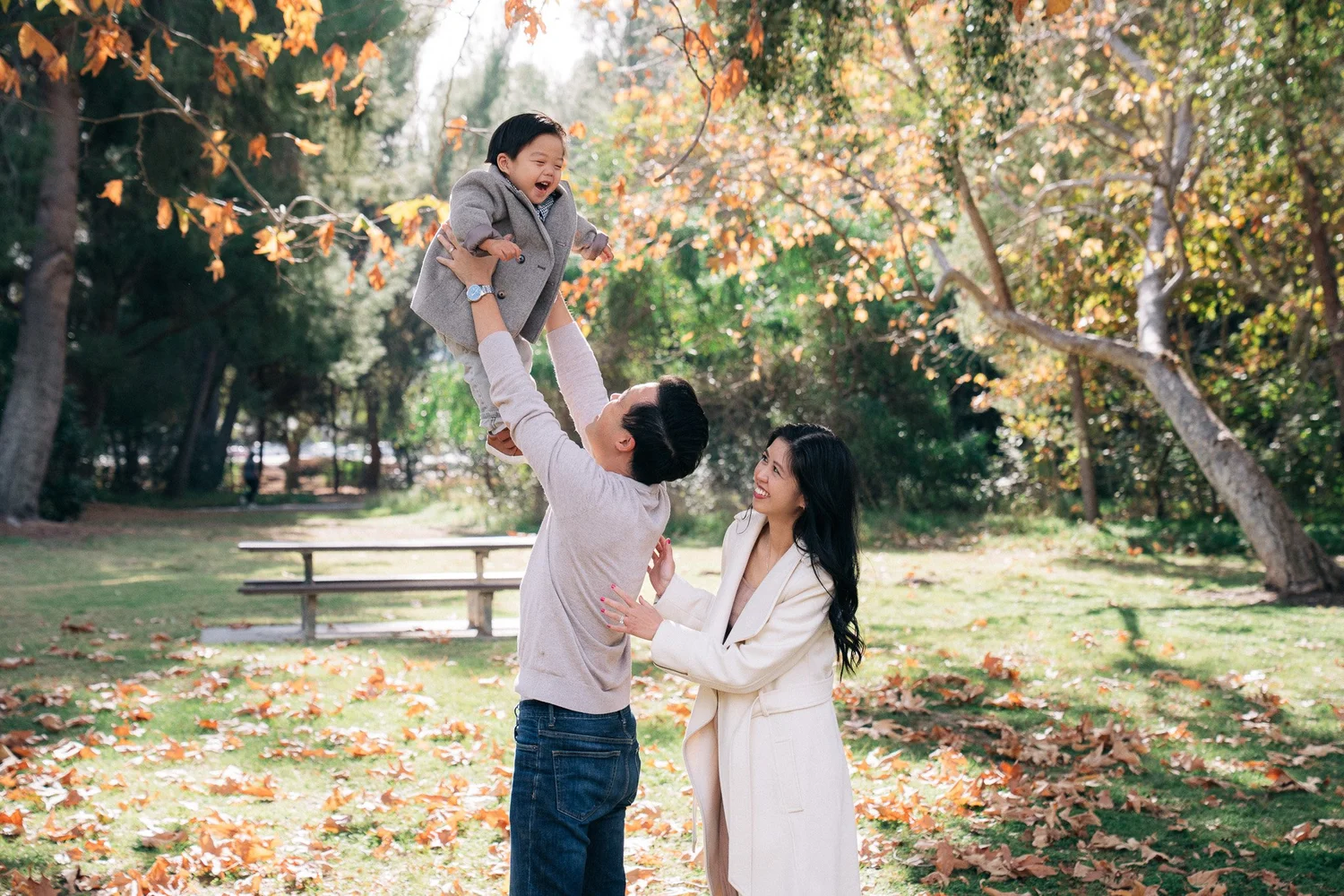 Father lifting his laughing toddler in the air beneath a canopy of golden fall leaves at an Orange County park