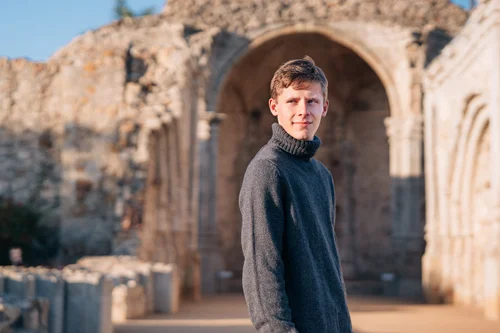A senior in a grey sweater in front of the stone arches of the Great Stone Church ruins at Mission San Juan Capistrano