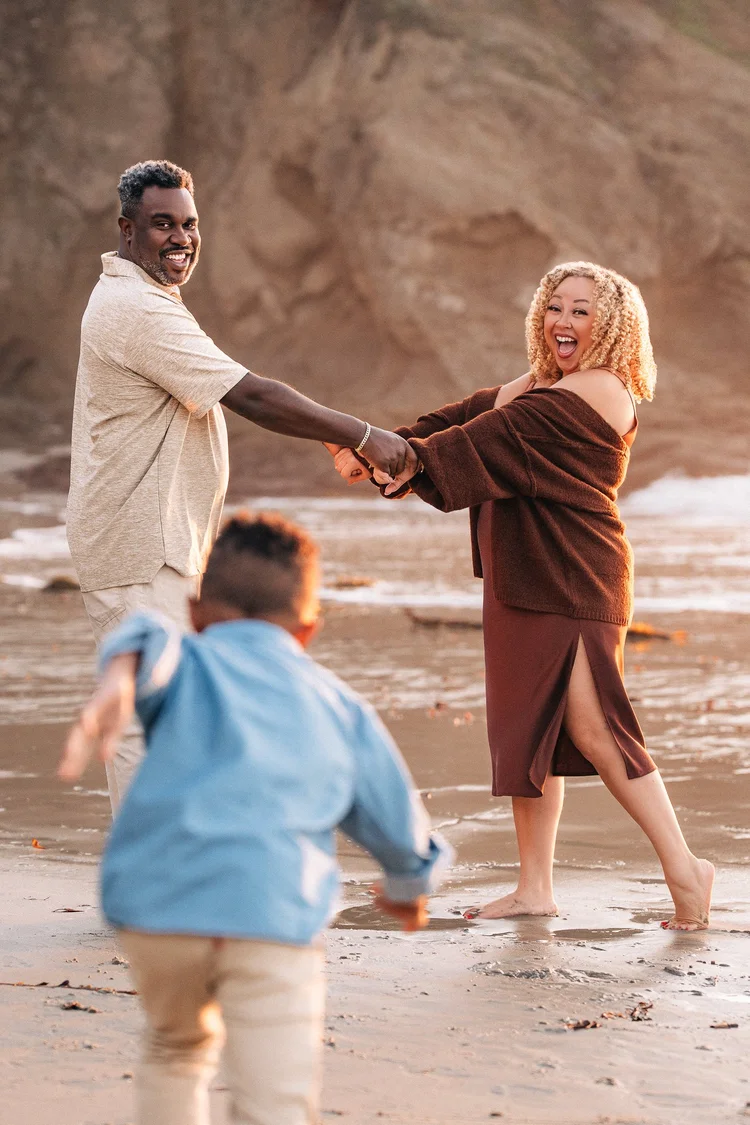 Parents holding hands and laughing on the beach while their young son runs in toward them