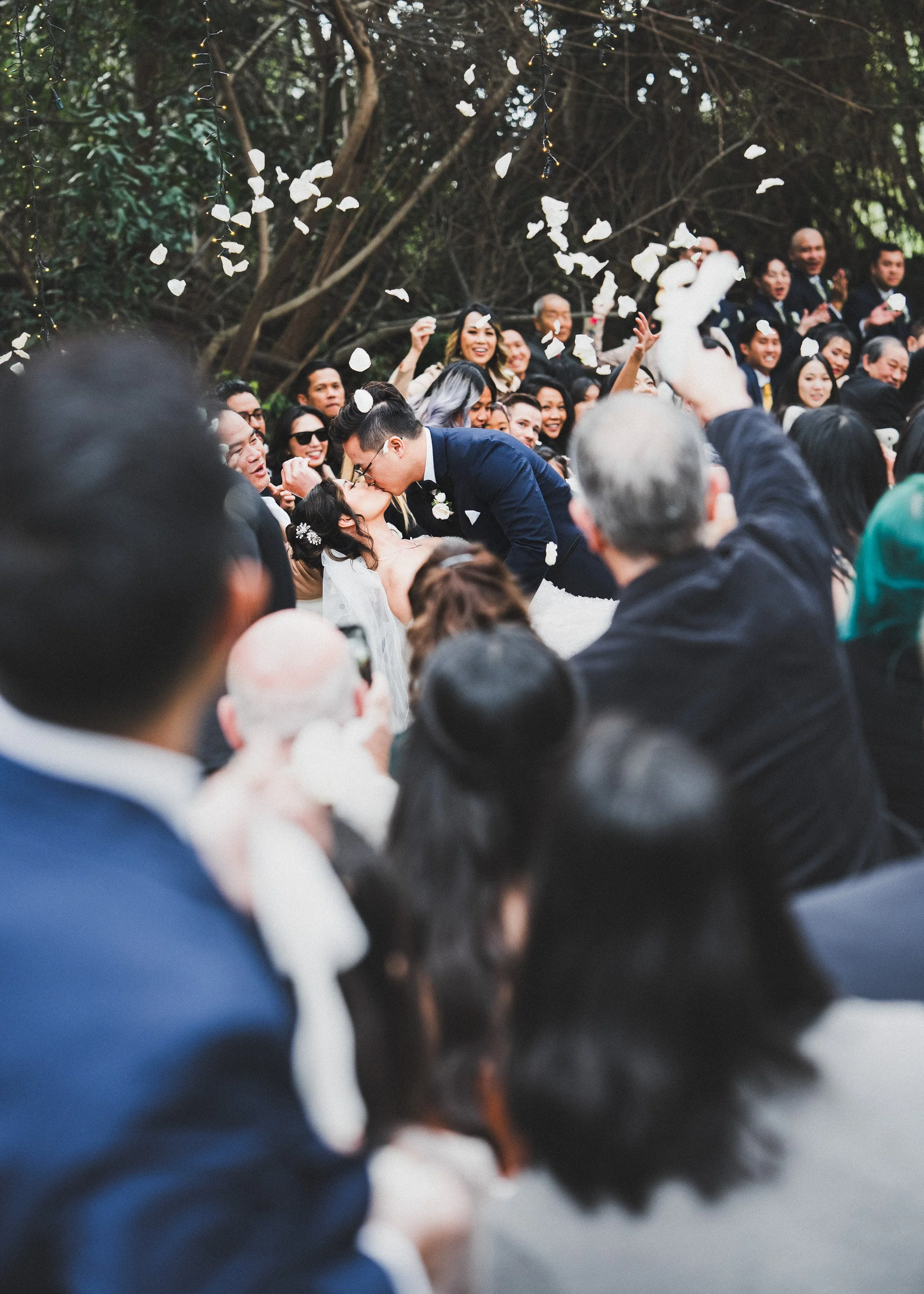 A bride and groom kissing in the middle of a cheering crowd while guests throw white petals under the trees