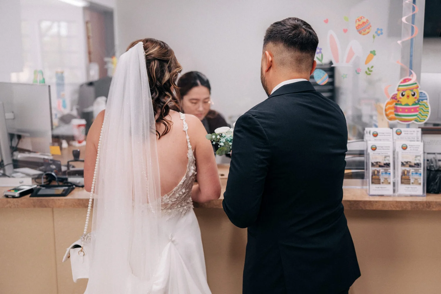 Jessica in her veil and Alex in his suit at the clerk's walk-up counter, seen from behind, with Easter decorations on the glass