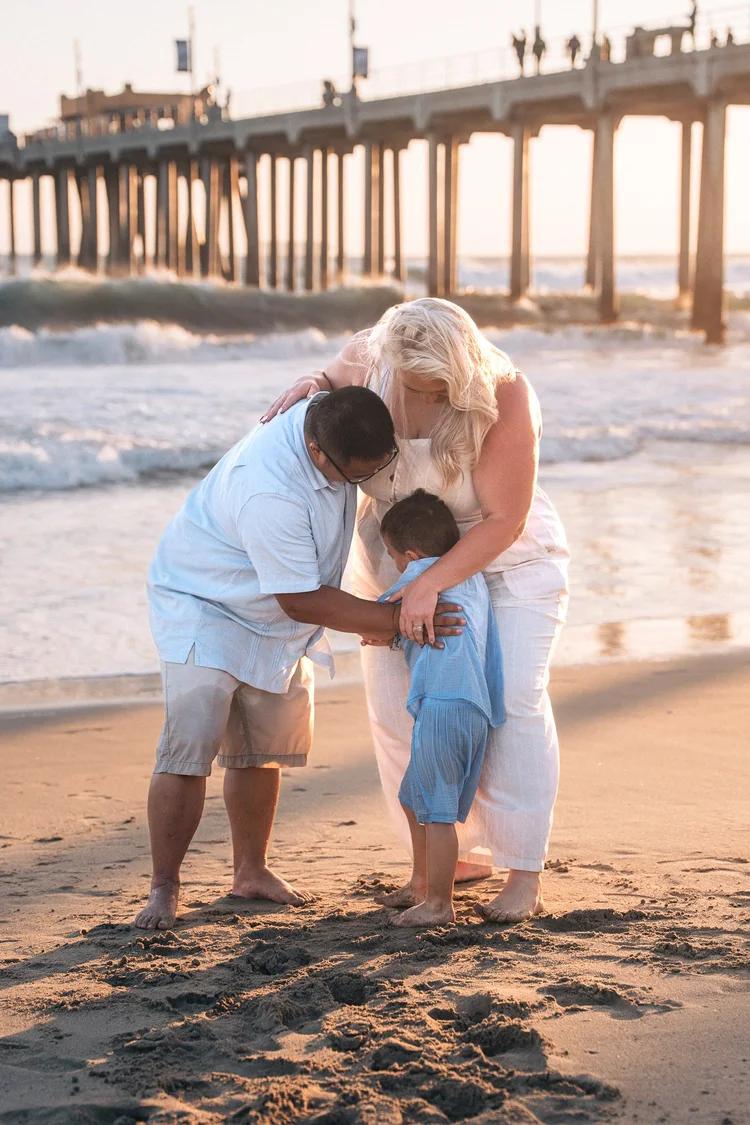 Both parents bent over their son in a hug at the edge of the surf, the pier behind them