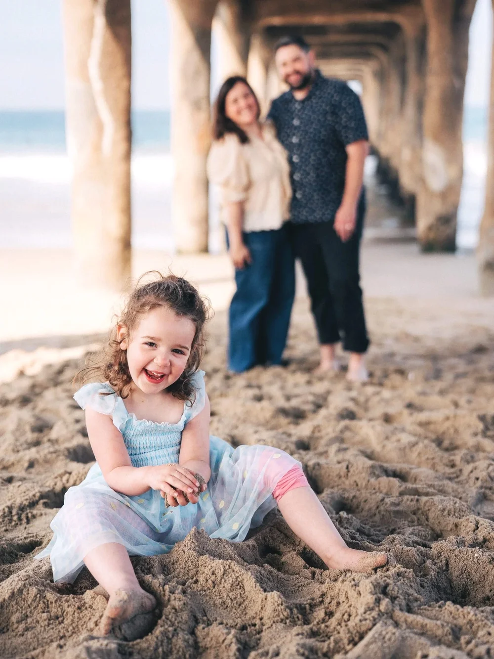 A child sitting in the sand under the pier smiling at the camera with her parents blurred behind