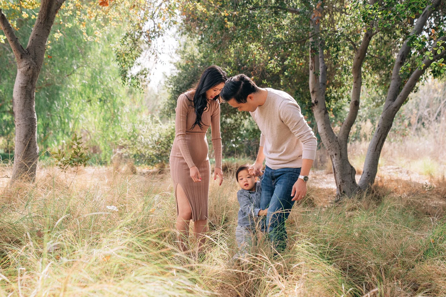A dad holding his young child's hand in tall grass while the child peeks shyly toward the camera
