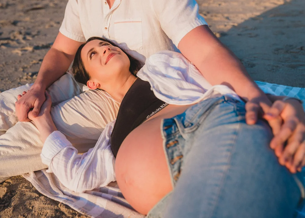 Mom lying back on a blanket on the sand at Strands Beach, looking up at her partner, their hands intertwined