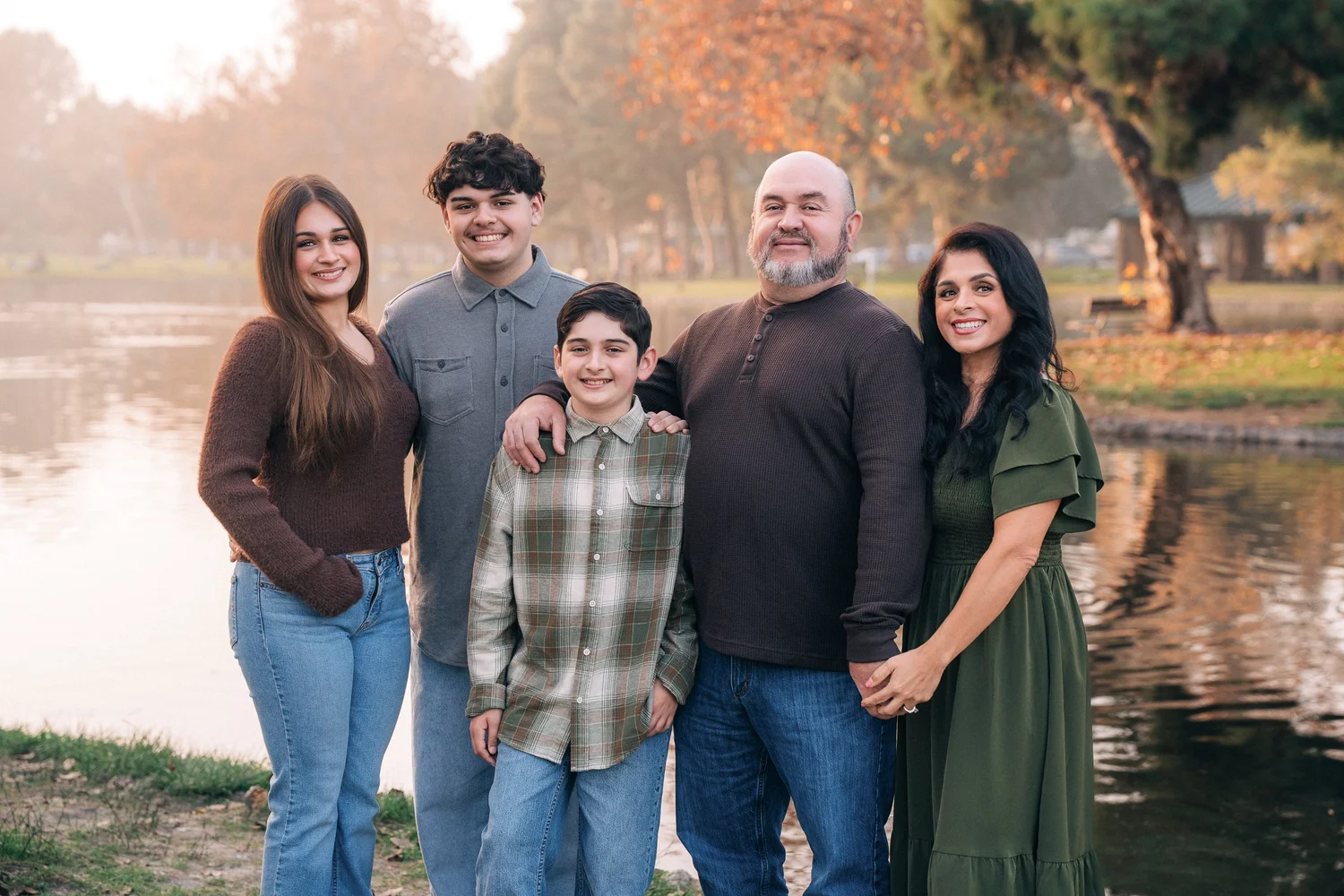 The Mirand family standing together by the lake at Yorba Regional Park, autumn trees behind them