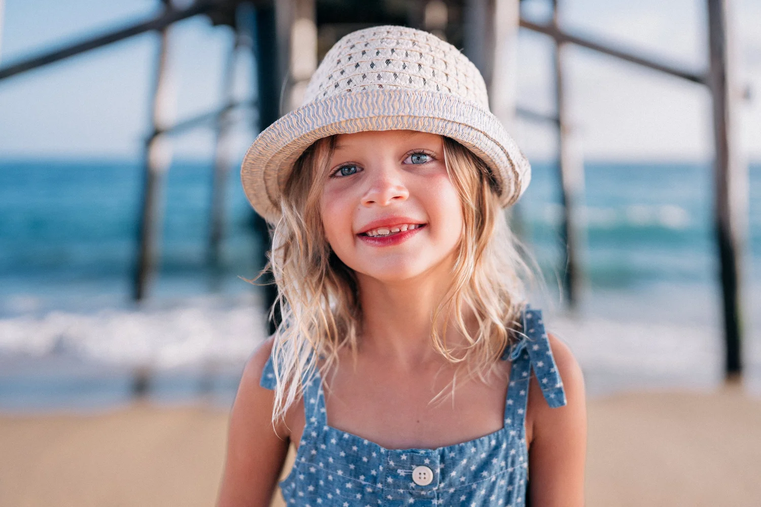 A young girl in a straw hat and chambray dress on the sand beneath the Balboa Pier in Newport Beach