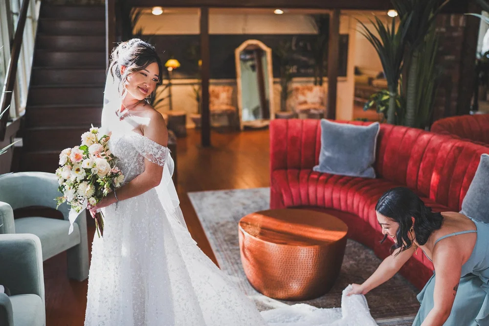 A bride holding her bouquet looks back over her shoulder while a bridesmaid in blue arranges her train, in a room with a red velvet sofa