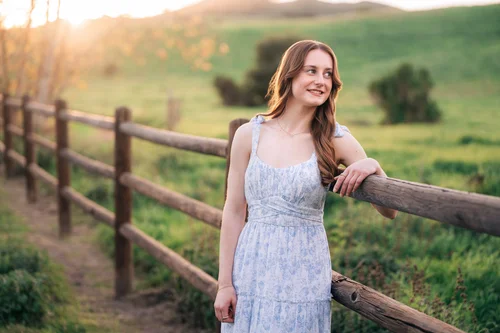 A senior in a floral dress leaning on a wooden fence in the green hills at Thomas F. Riley Wilderness Park