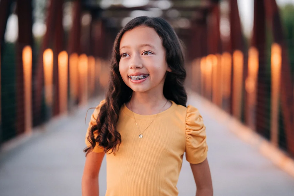 The younger Slemmons daughter in a yellow top smiling on the bridge, the railings glowing warm behind her