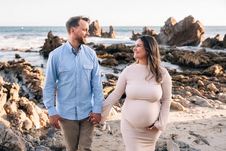 An expecting couple holding hands on the rocks at Little Corona del Mar in Newport Beach