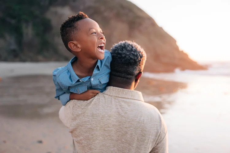 A boy laughing over his dad's shoulder on the sand at Strands Beach in Dana Point