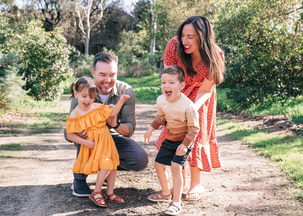 The whole Leon family mid-laugh on the path, both kids wriggling out of their parents' arms