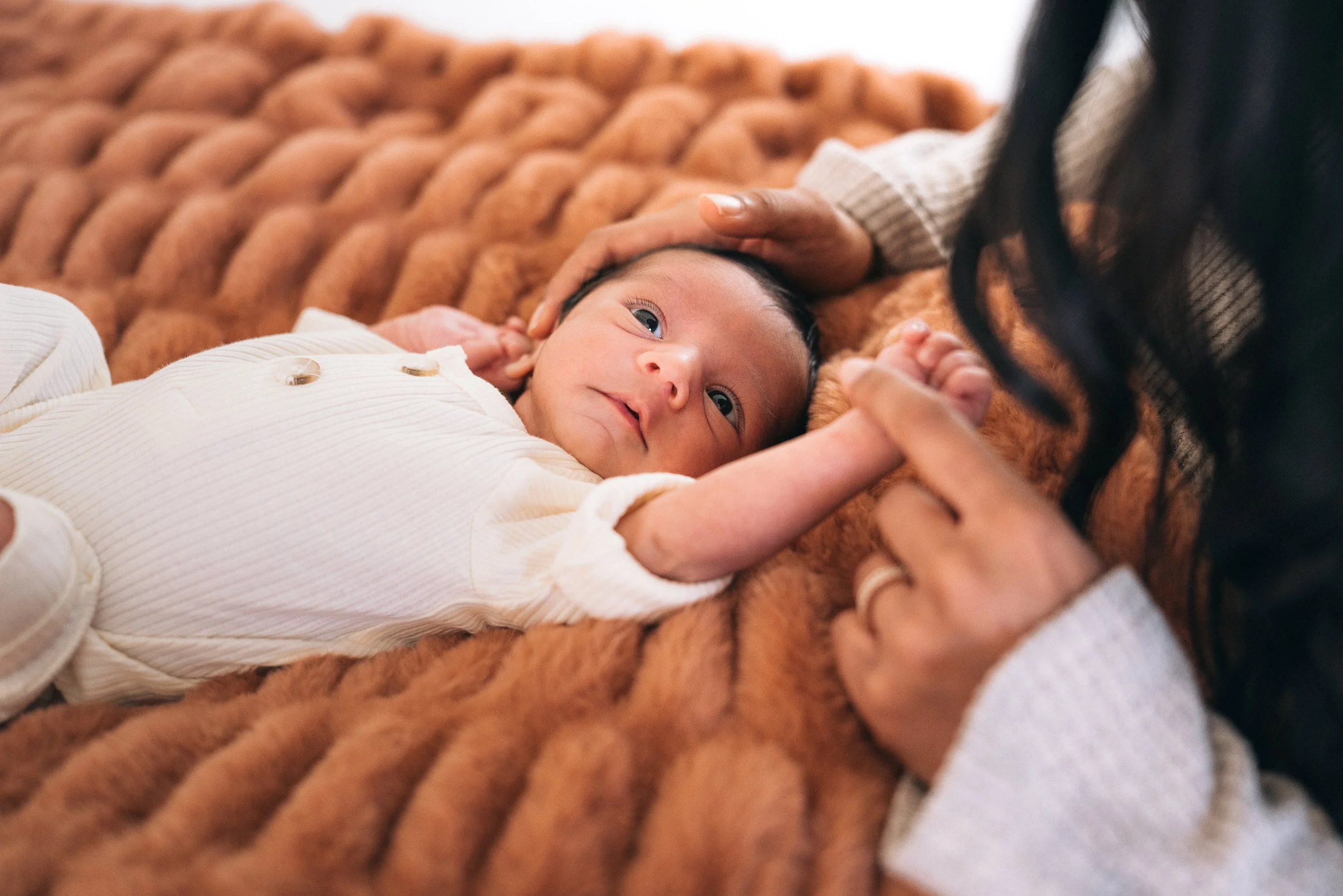 Newborn in a white onesie resting on a rust knit blanket with a parent's hand on her head, in-home newborn session in Orange County