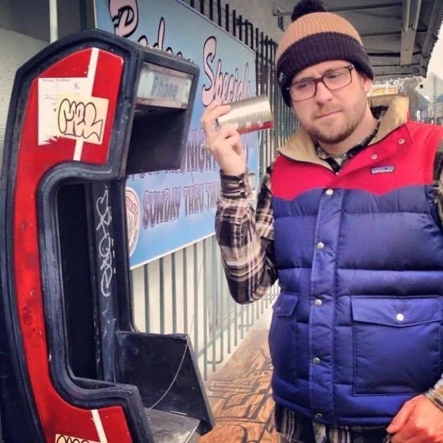 Daniel Wesley at an old pay phone, holding a tin can on a string to his ear where the handset used to be