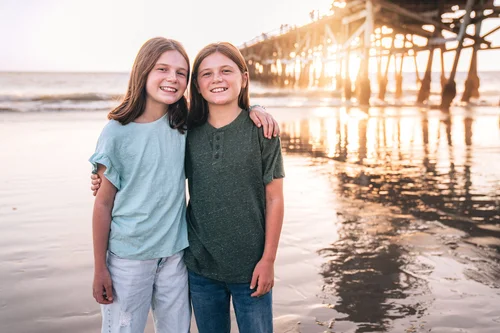 Two siblings arm in arm on the wet sand at sunset with the San Clemente Pier glowing behind them