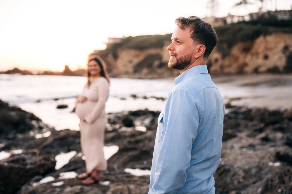 Dad in the foreground looking out toward the water at Little Corona del Mar, mom soft in the background with both hands on her bump