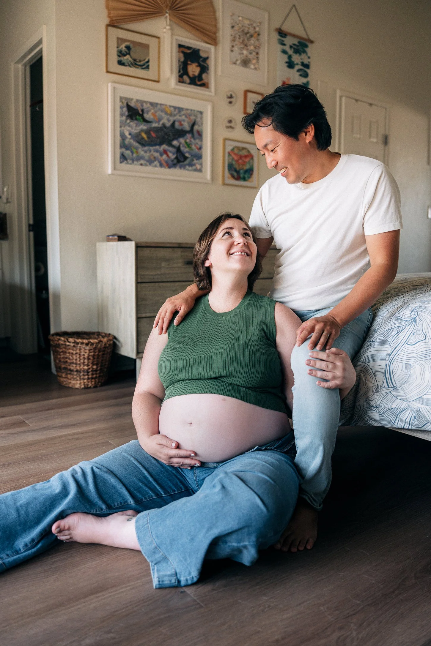 In home maternity session, the mother in jeans and a green top with her partner in light jeans and a white tee