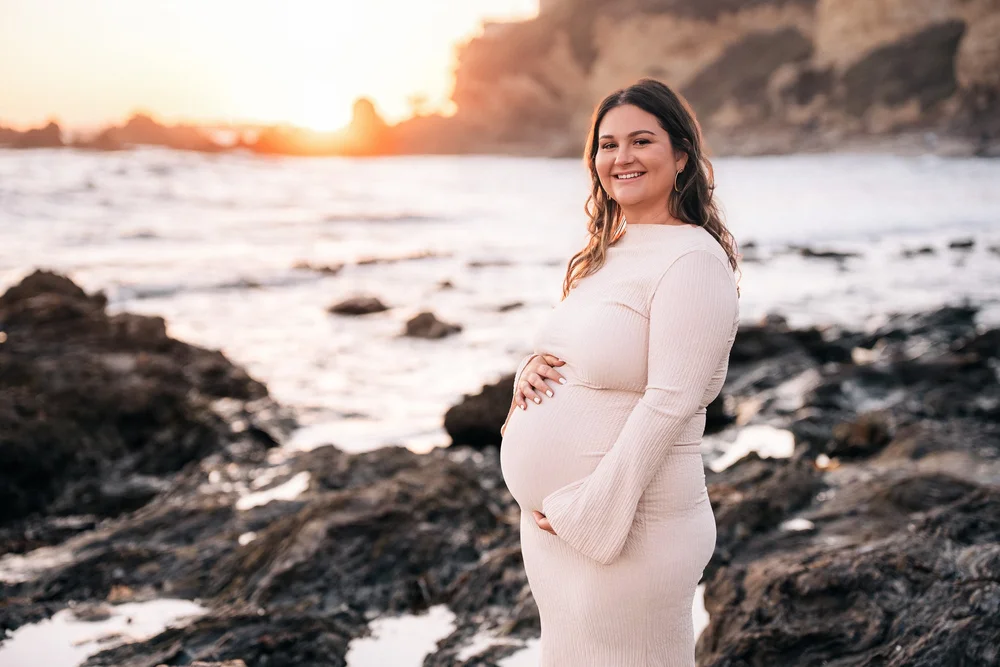 Mom in a cream dress on the rocks at Little Corona del Mar, both hands on her bump, the sun setting behind the sea stacks
