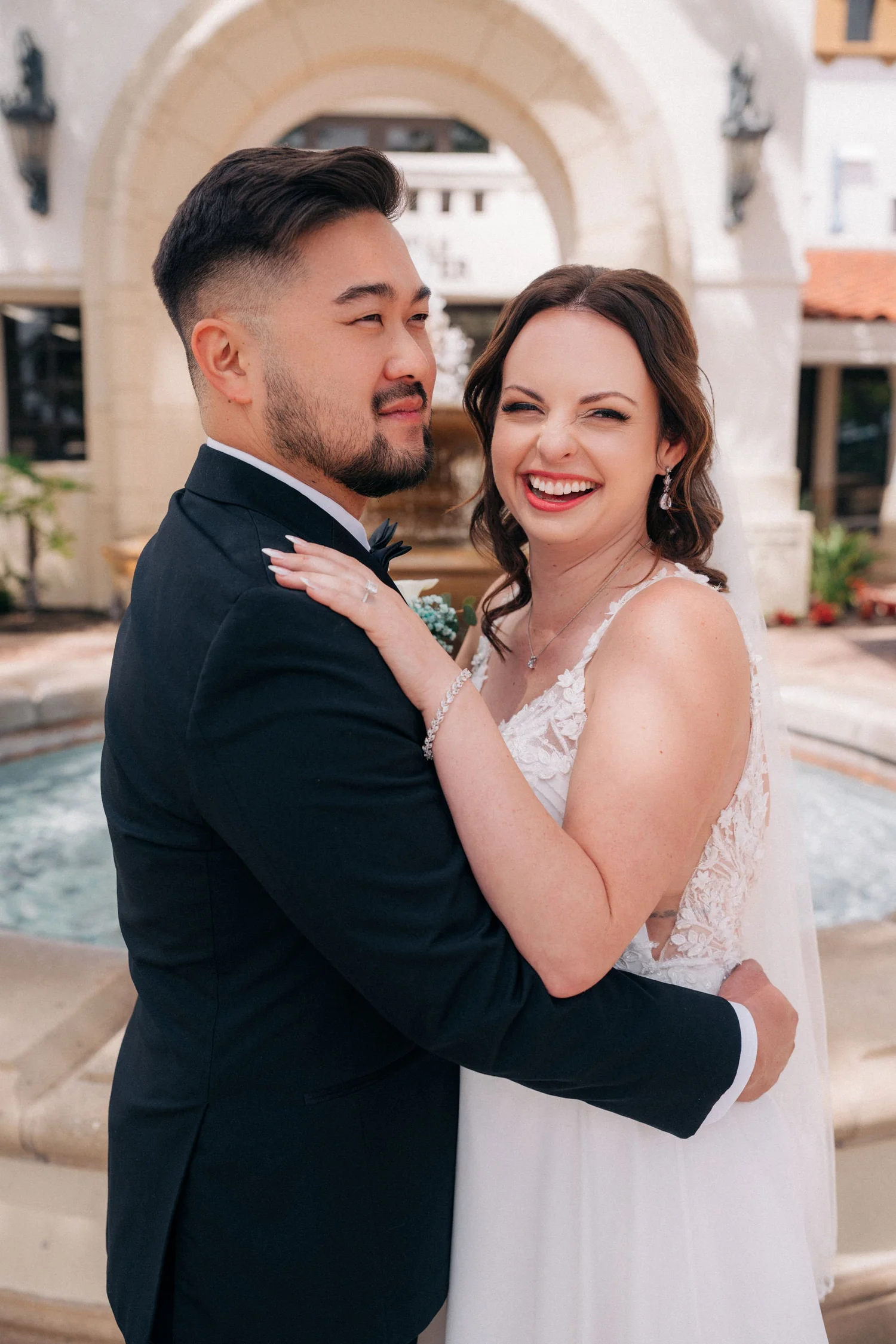 Jessica laughing while Alex holds her, in front of an arched courtyard with a fountain after their courthouse wedding in Laguna Hills