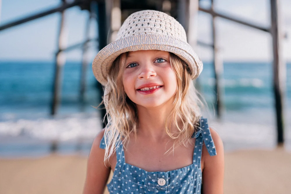 Little girl in a woven sun hat smiling under the pier, candid Orange County family photos