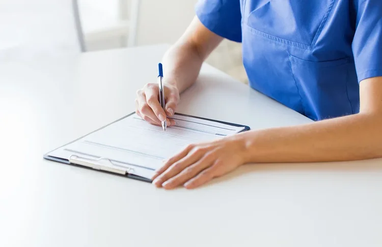 A healthcare professional in blue scrubs writing on a clipboard