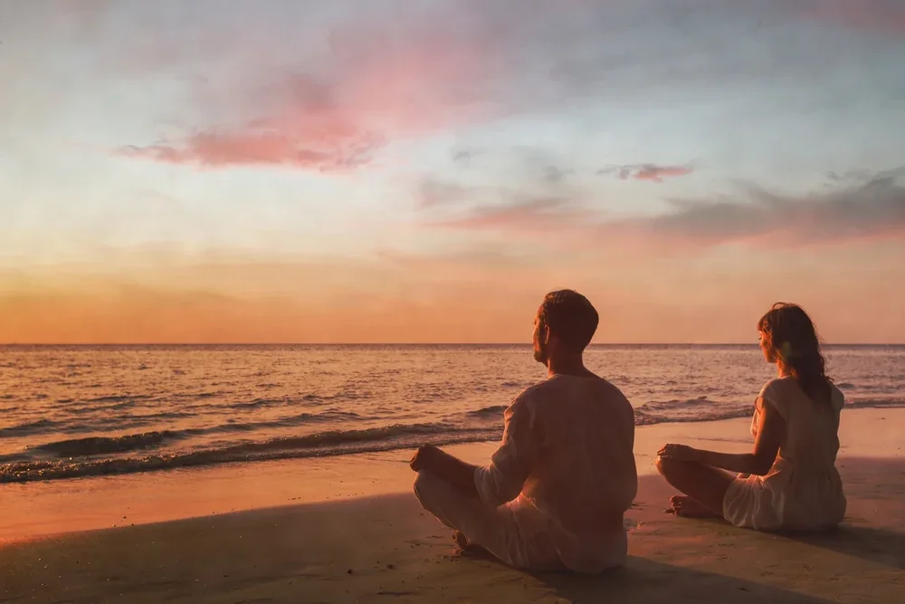 A man and woman meditating together by the water at sunset