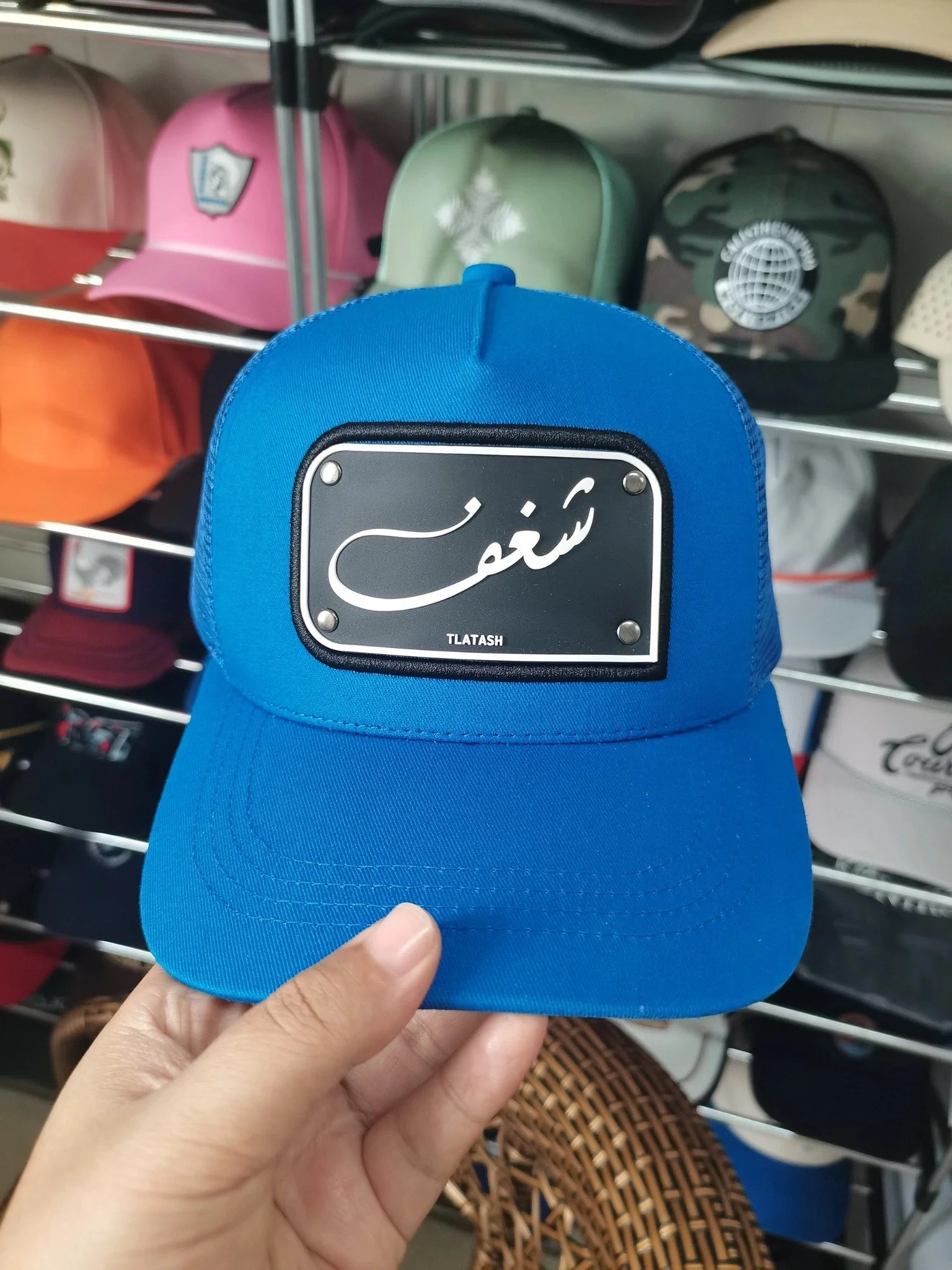 Blue cap sample with a rectangular black front badge, white lettering, and four silver-tone studs
