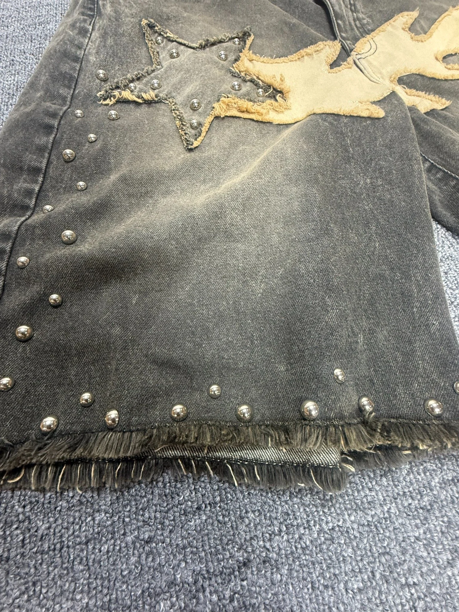 Gray denim sample detail with star appliqué, tan fabric shapes, silver-tone studs, and frayed edges