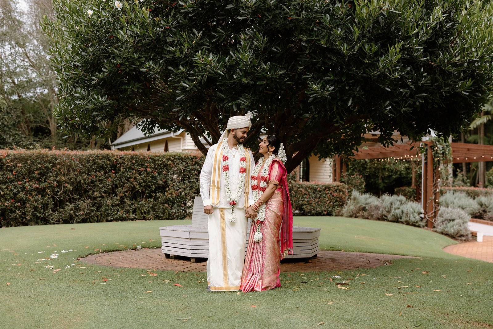 A couple in traditional wedding attire beneath a garden tree at Shambala Estate.