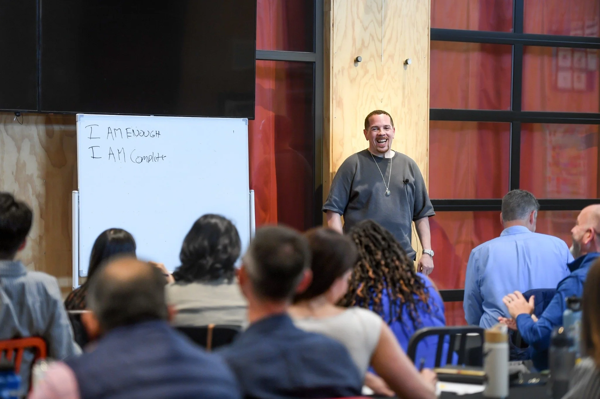 Sean Goode smiling while facilitating a seated group, beside a whiteboard reading “I am enough” and “I am complete.”