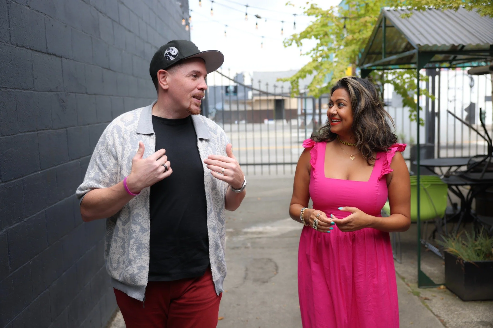 Two people walk outdoors in conversation, one gesturing as the other smiles and listens.