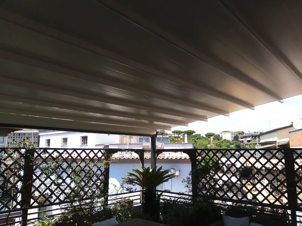 Un giardino pensile, con vista su Roma, ricco di piante rampicanti su pannelli grigliati, completamente ombreggiato dalla nostra pergotenda inclinata realizzata in alluminio bianco e PVC.