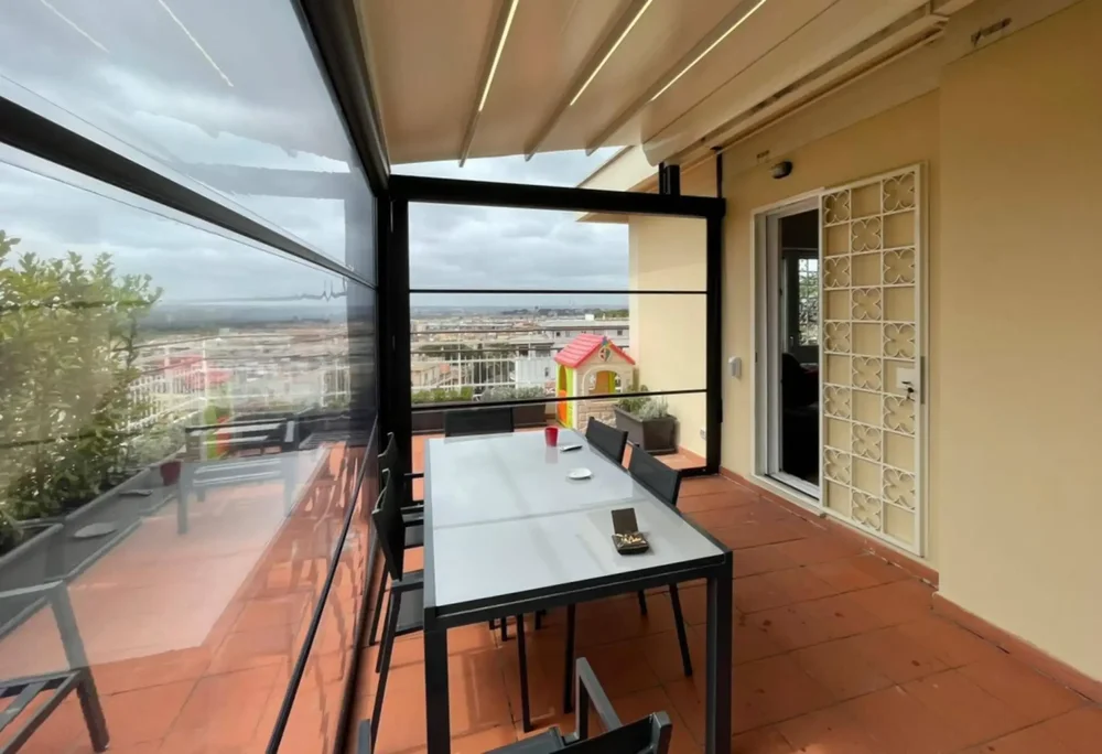Ampia terrazza panoramica protetta da chiusure laterali antivento in PVC Cristal, con vista su Roma.