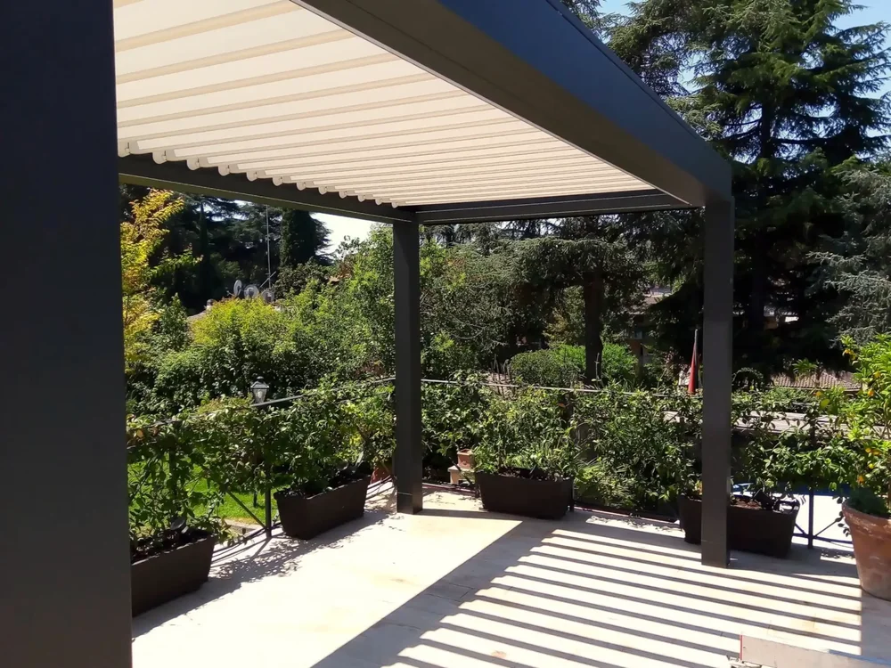 Pergola bioclimatica con struttura nera e lamelle in alluminio bianche, completamente aperta per far passare il sole, che può essere chiusa elettronicamente quando le condizioni meteorologiche lo richiedono.