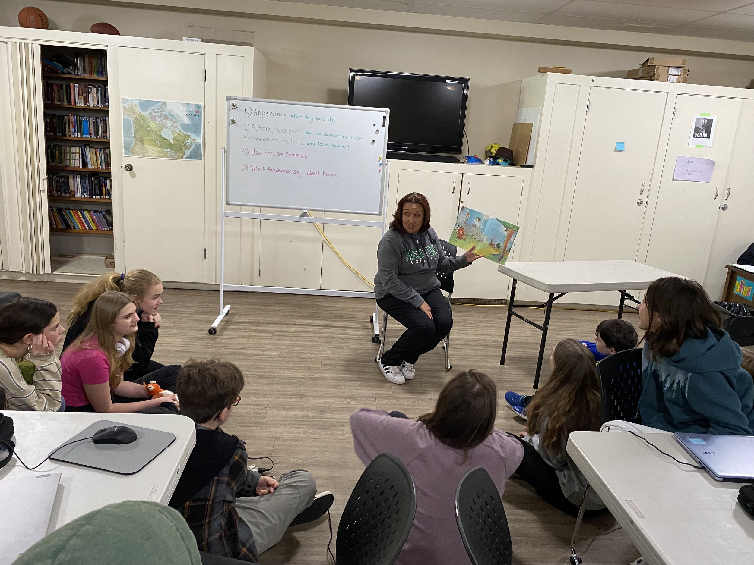 Students sit around the teacher listening while she reads a picture book to them