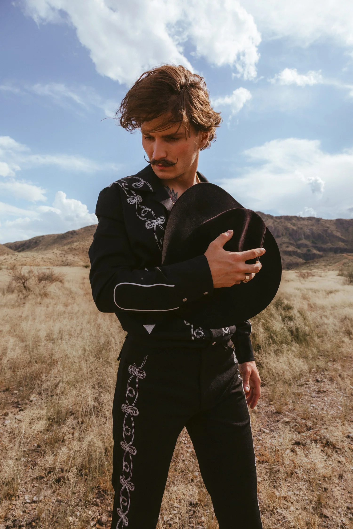 Tru Western Leather No. 5 Canyon Echoes campaign, Braxton Keith in a black embroidered western suit holding his hat against his chest in the West Texas desert, mountains and clouds behind