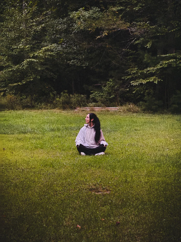 Laura sitting cross-legged in a mown field with a treeline behind her