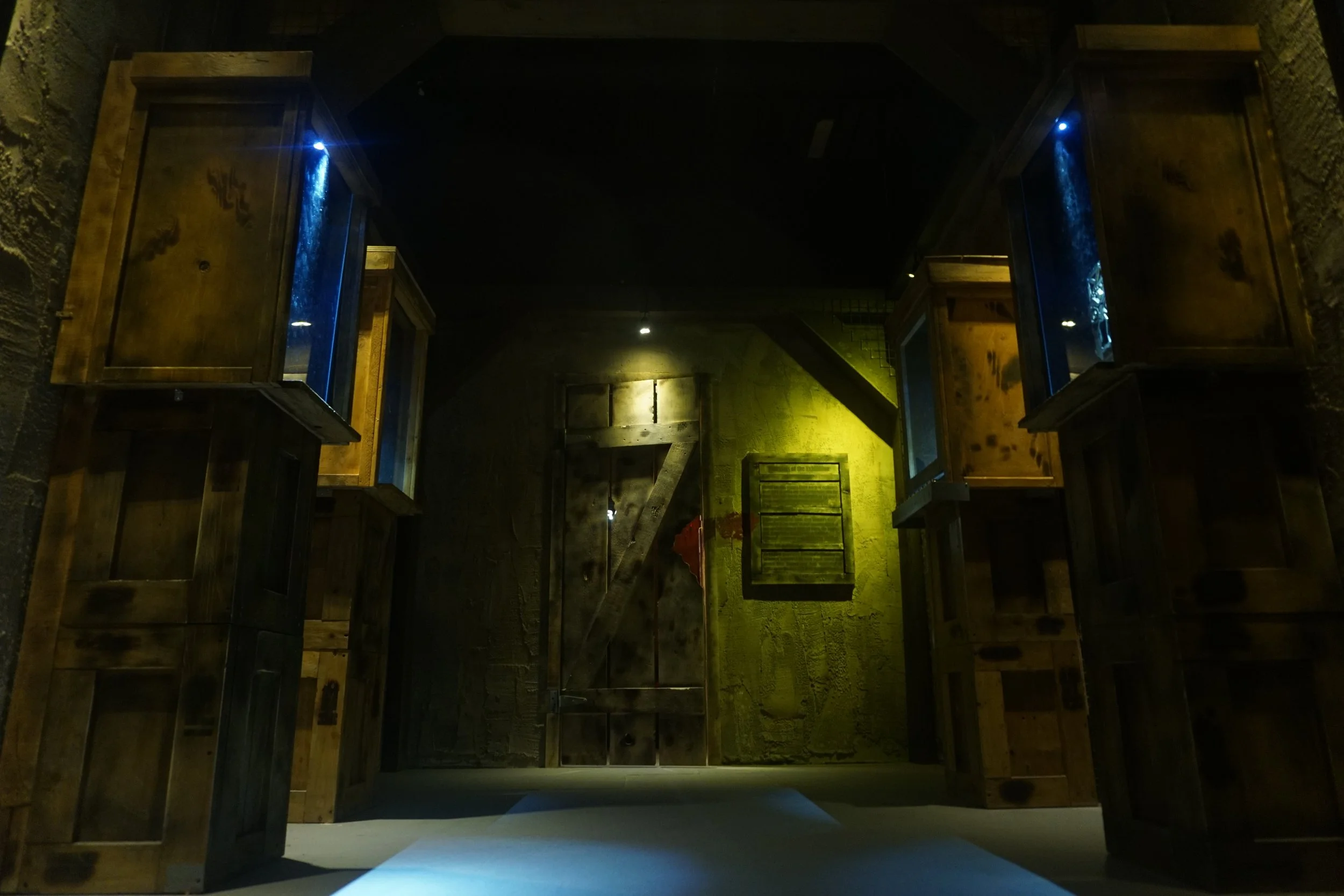 Industrial stone hallway with stacked crates and blue-lit display cases leading to a heavy door.